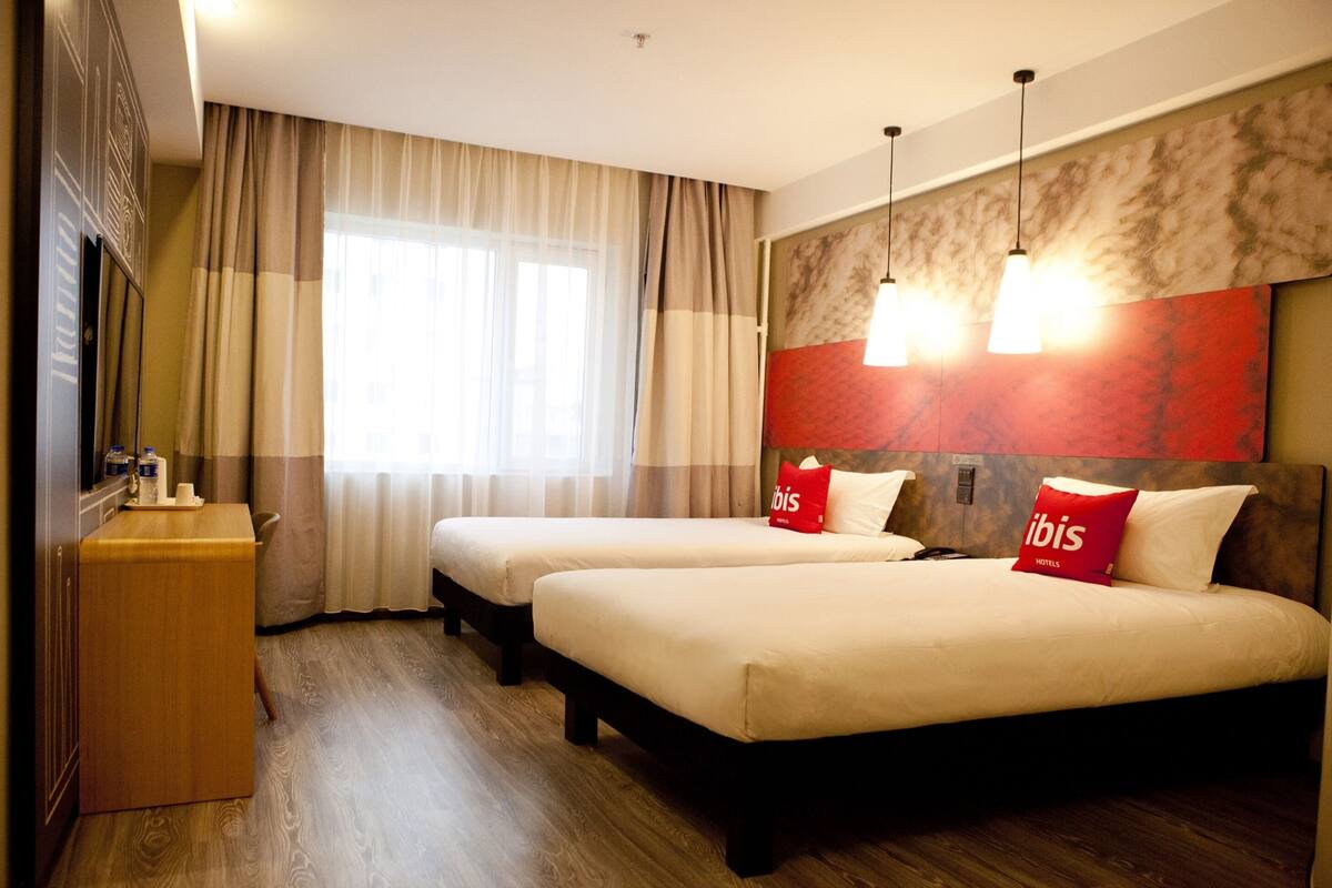 A hotel room with two beds, a wooden desk, and a large window with curtains.