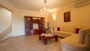Un salon avec un canapé, une table basse et un lustre. Un escalier se trouve à gauche, ainsi qu'un meuble avec des portes en verre.