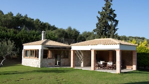 Une maison en pierre à deux étages, avec un toit en tuiles, une terrasse dotée de mobilier d’extérieur et une vue sur une colline boisée.