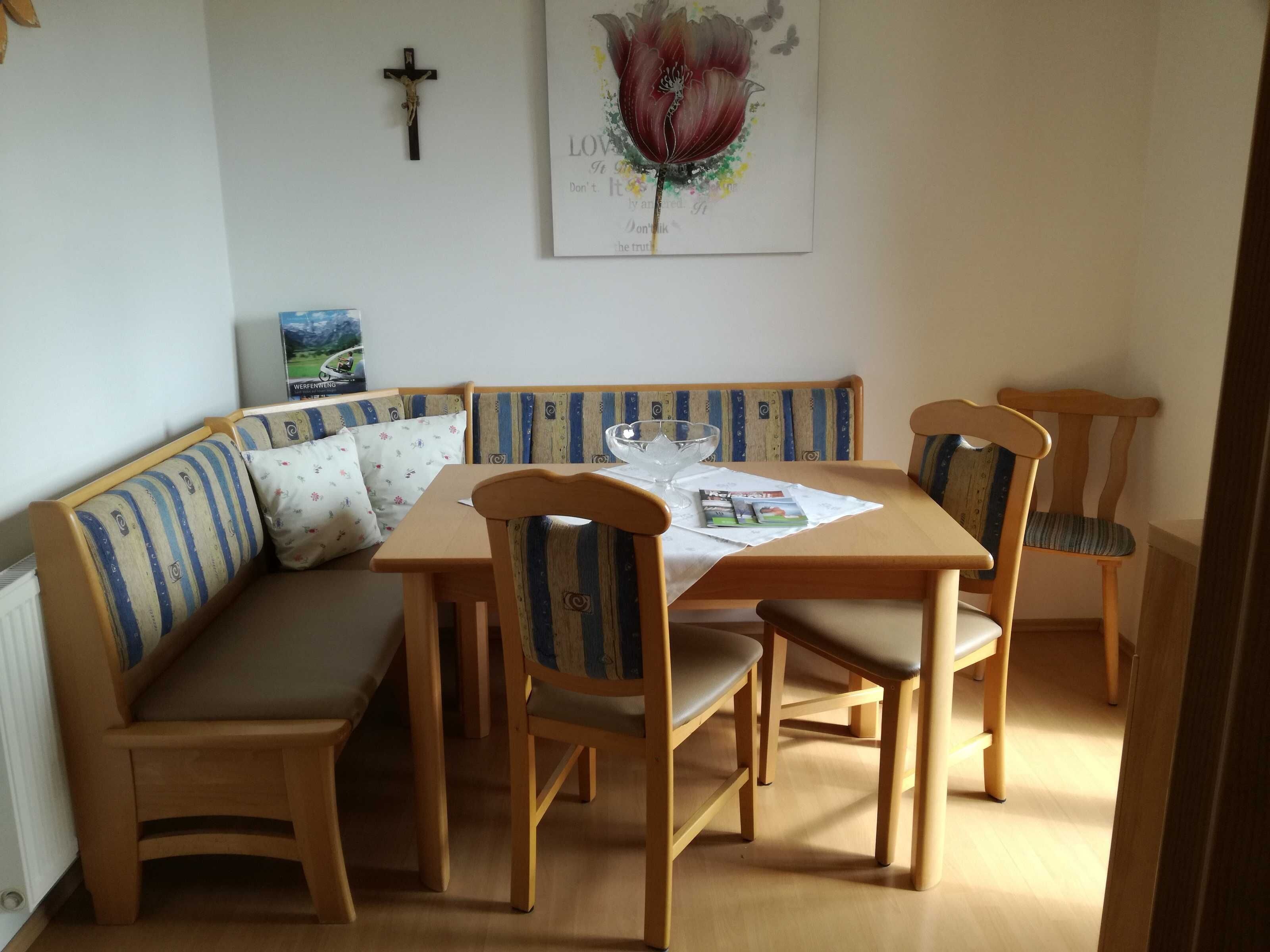 Een eetruimte met een houten tafel, vier stoelen, een bank met gestreepte kussens, en een aan de muur bevestigd kruis.
