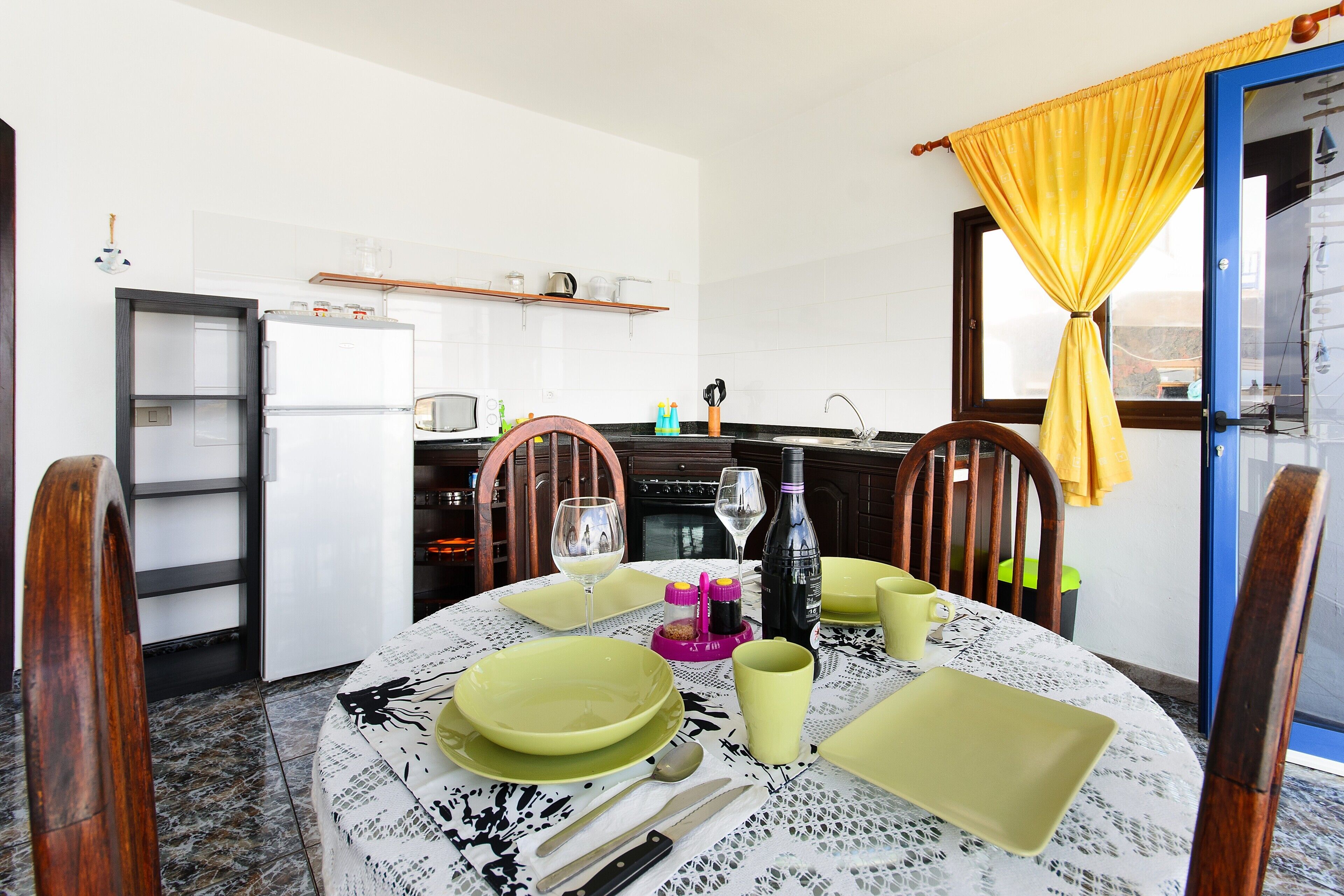 Un coin repas avec une table ronde dressée pour quatre personnes, une cuisine en arrière-plan et une fenêtre ornée de rideaux jaunes.