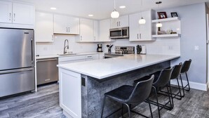 A modern kitchen with white cabinets, stainless steel appliances, a marble island, and black bar stools. - Scorpio #104 2 Bedroom Condo by RedAwning (Vail)
