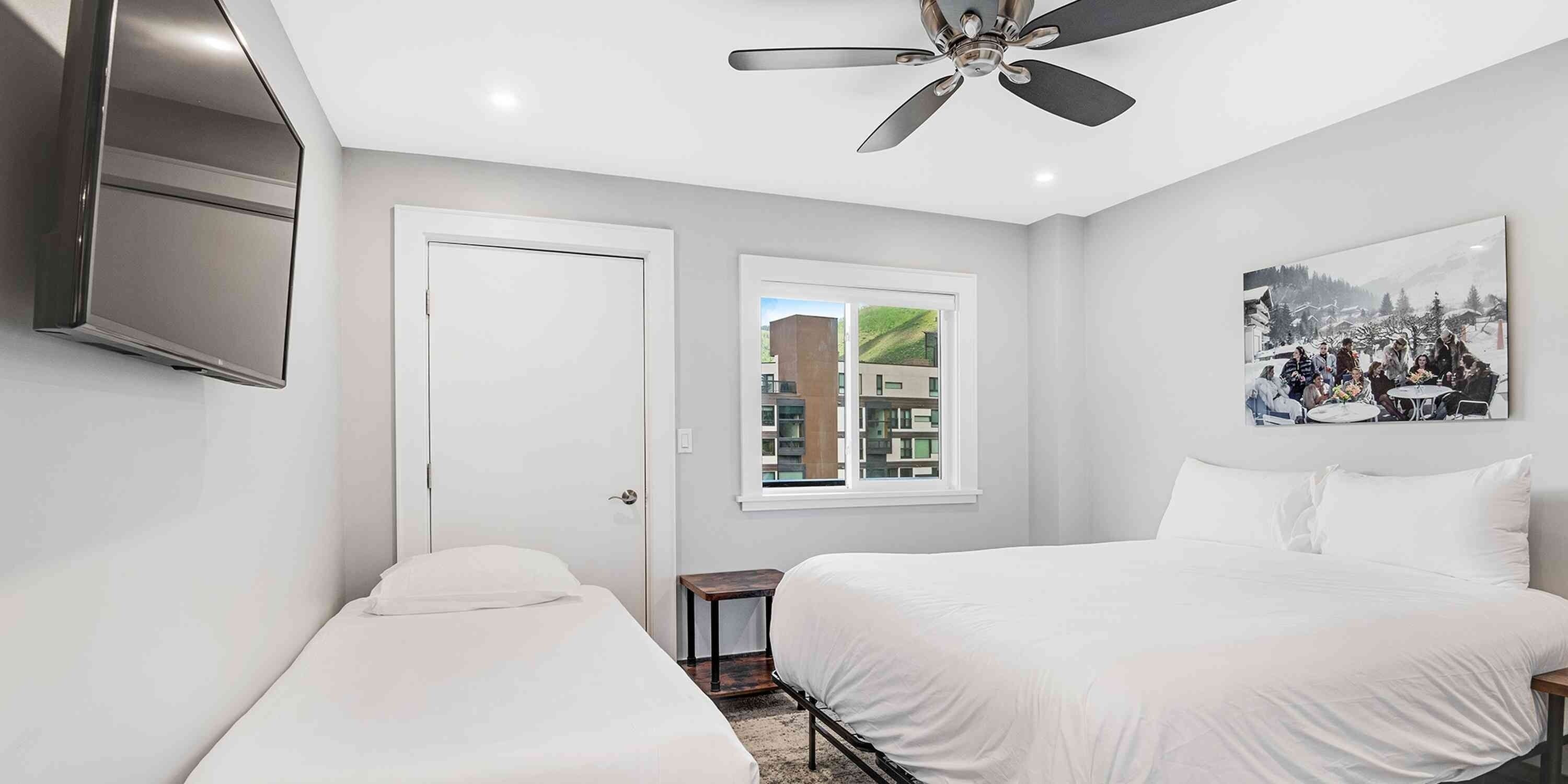 A bedroom with two beds, a ceiling fan, a TV, a window with a view of buildings, and a painting on the wall.