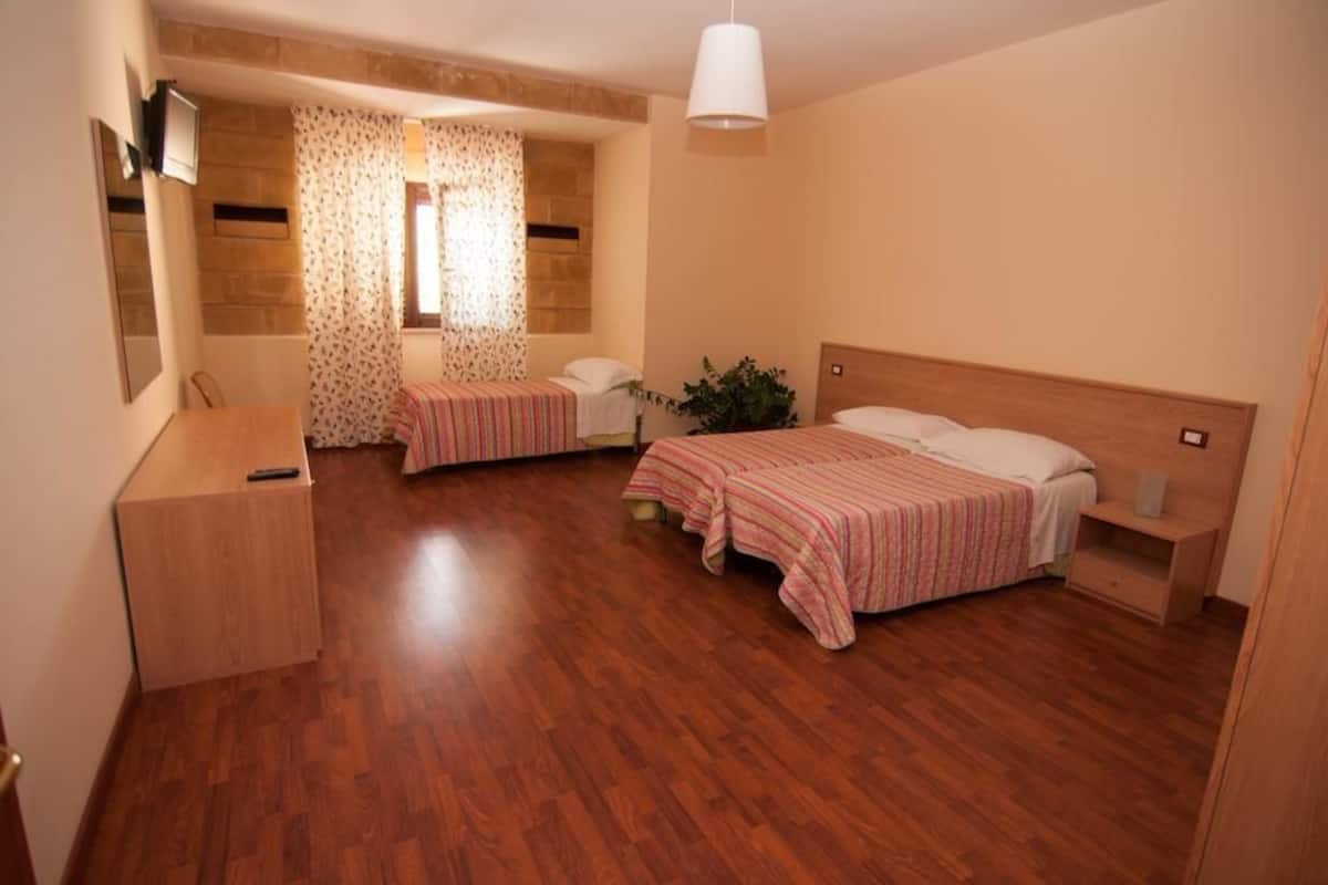 A room with two beds, wooden flooring, a desk, a mirror, and a window with patterned curtains.