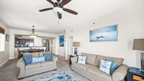 A spacious living room with a beige sofa set, a blue and white patterned rug, a ceiling fan, and a painting of a seascape. - Destin Miramar Sweeping Ocean Views ~ Five Swimming Pools ~ Relaxing! End Unit.  (Destin)