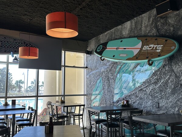 A restaurant interior with a surfboard mural, large windows, and modern furnishings.