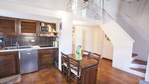 A kitchen with wooden cabinets, a refrigerator, and a microwave. There is a dining table with chairs and a staircase leading to an upper level. - Appartamento La Perla (Marciana Marina)