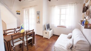 A cozy room with a wooden dining table, chairs, a small TV on a white cabinet, a white sofa, and a window with curtains. - Appartamento La Perla (Marciana Marina)