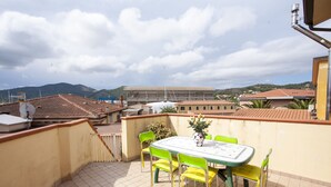 A rooftop terrace with a table and chairs, a potted plant, and a view of buildings and mountains in the background. - Appartamento Trilo Ornella (Portoferraio)