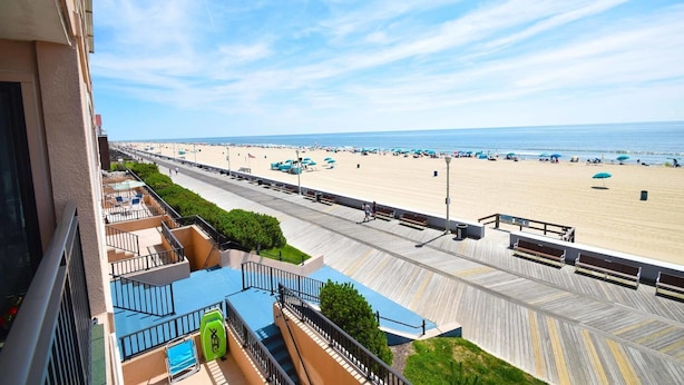Uitzicht op een strand met een houten promenade, een zwembad en strandparasols vanaf een balkon.