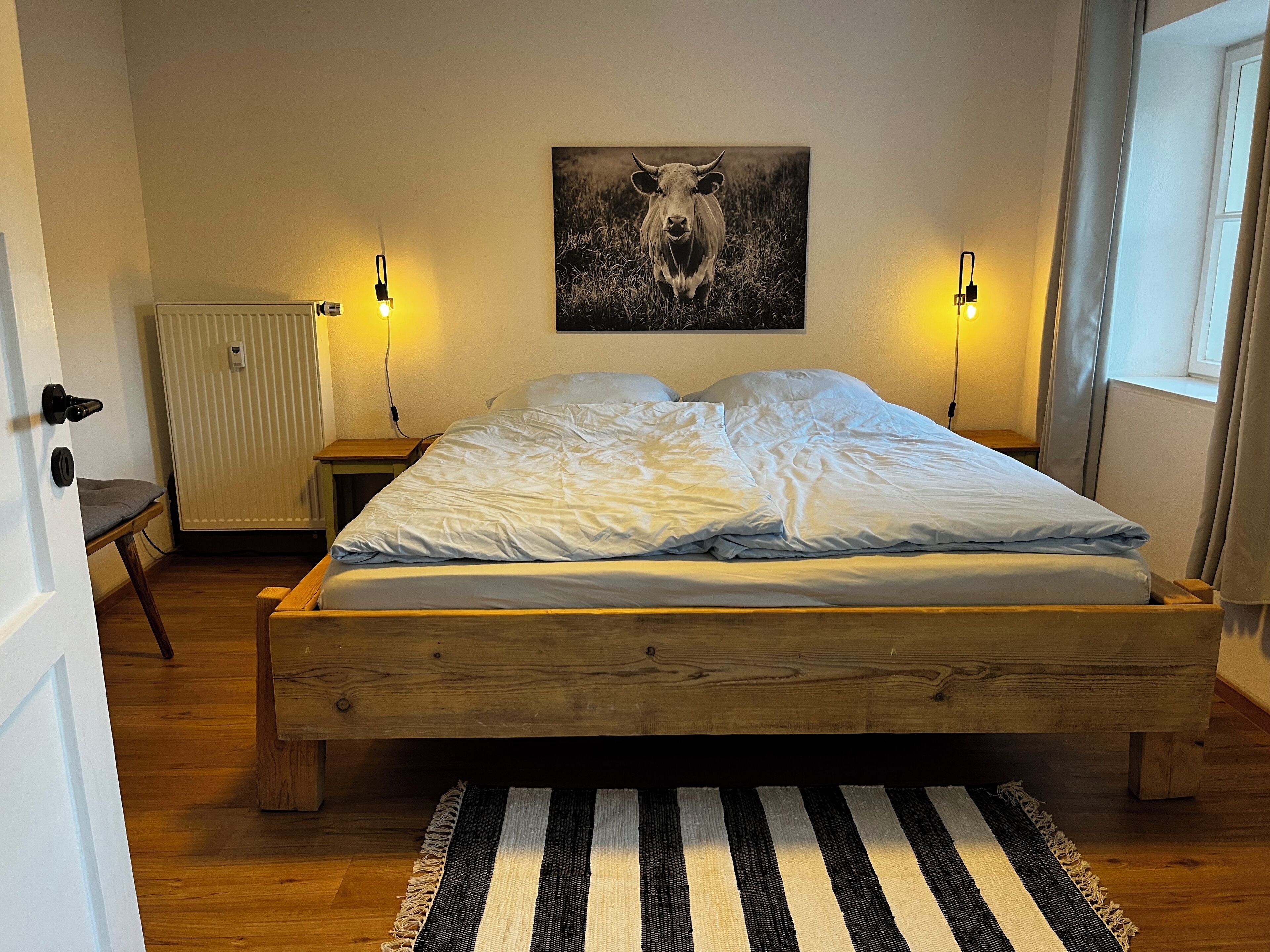 Een slaapkamer met een houten bed, twee bedlampen, een radiator en een zwart-wit streepig vloerkleed.