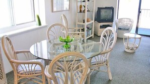 A dining area with a glass table surrounded by wicker chairs, a television, and a bookshelf. - Even Keel 404 2 Bedroom Condo by RedAwning (Ocean City)