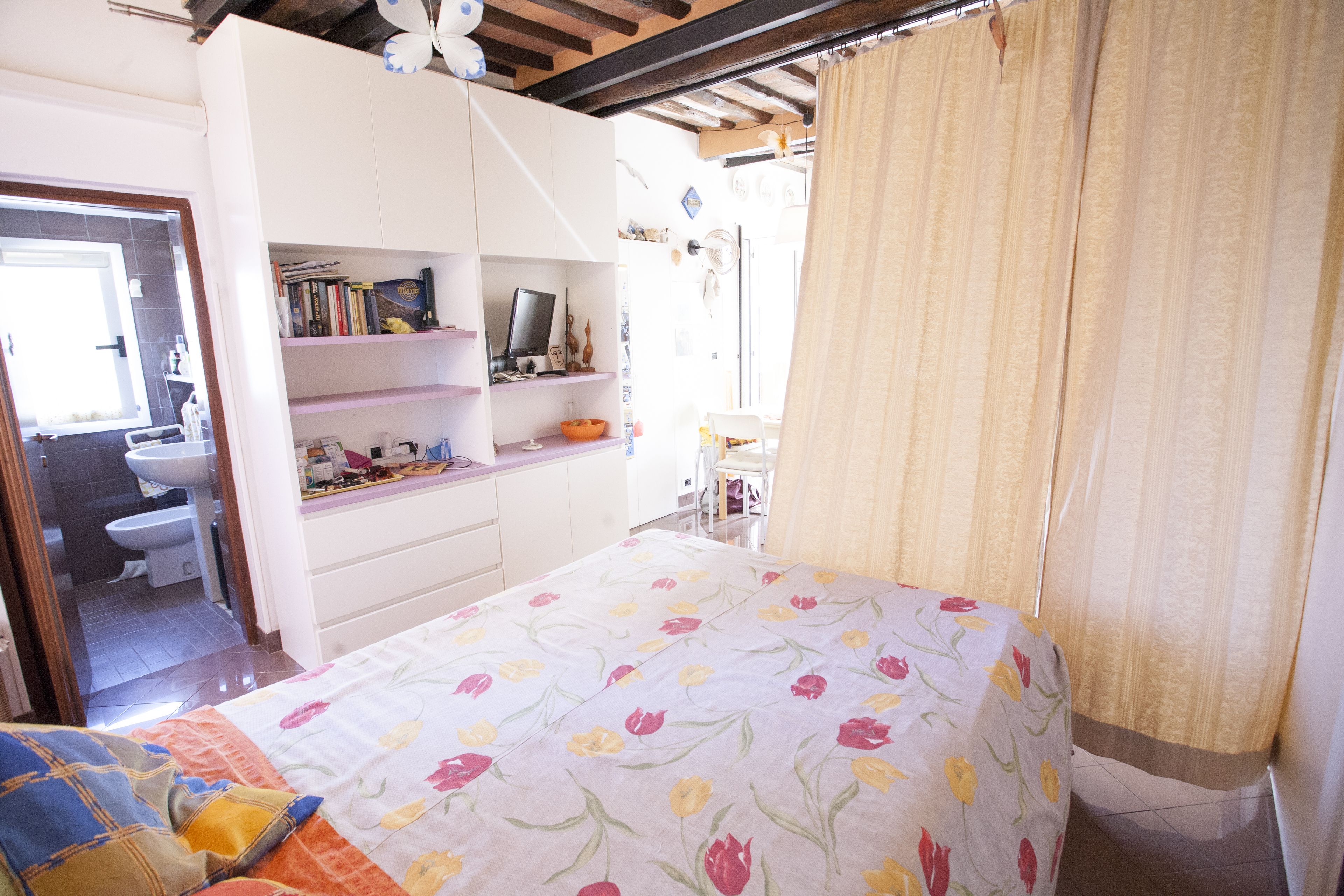 Ein Schlafzimmer mit Bett, Schreibtisch inklusive Computer, Bücherregalen und einem durchgehenden Türspalte sichtbaren Badezimmer.
