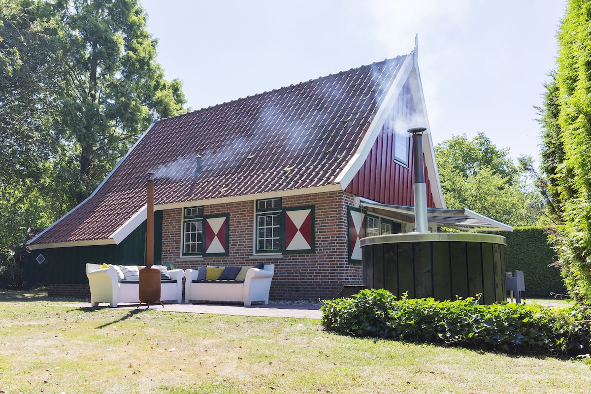 A house with a sloped roof, a chimney emitting smoke, and a garden area with a white outdoor sofa and a wooden table.