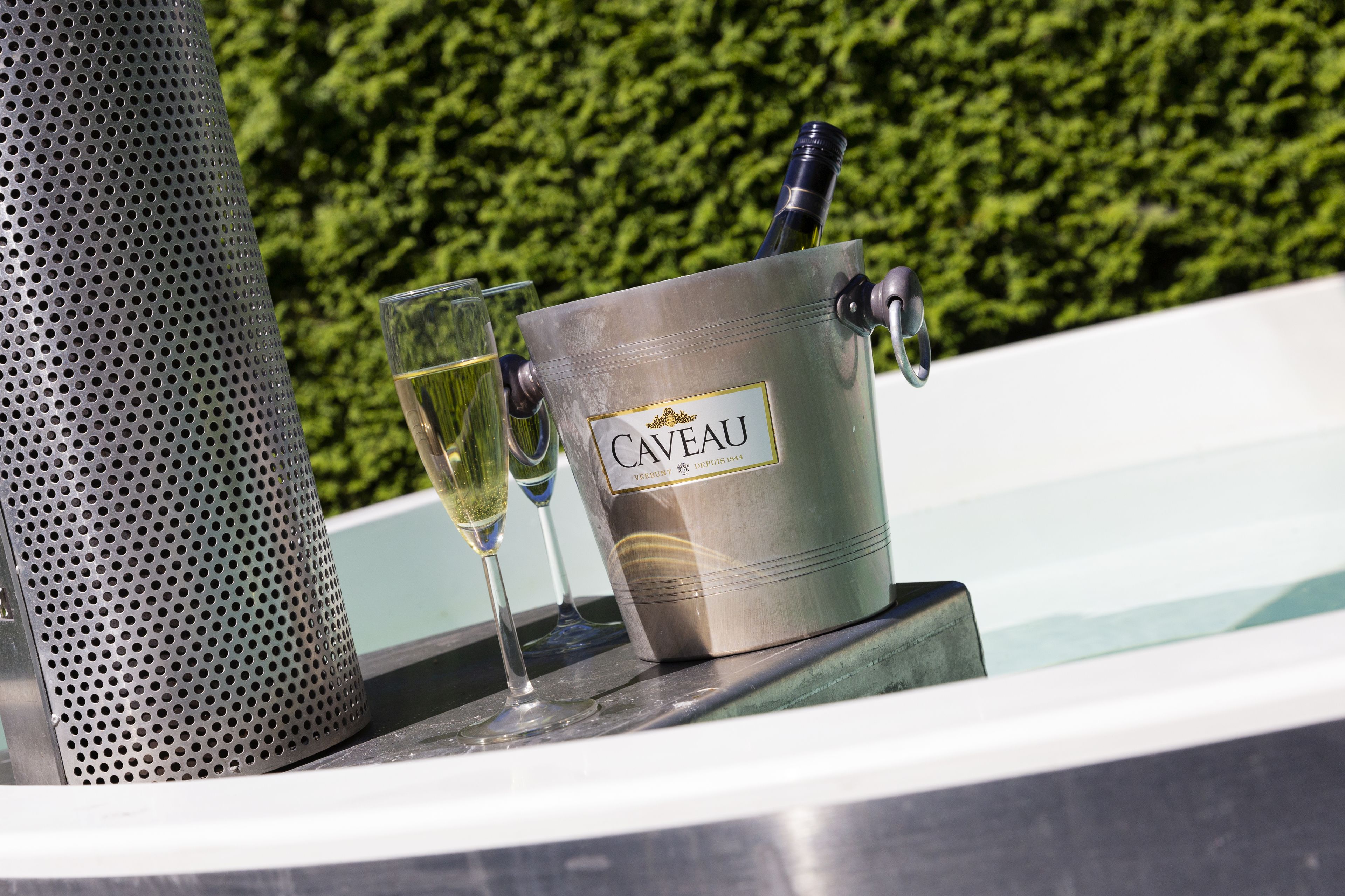 A metal bucket labeled "Caveau" with a bottle and glasses on a poolside.