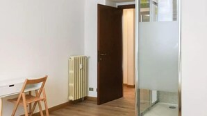 A room with a wooden chair, a radiator, a door, and a glass shower enclosure. - Corso Sempione (Milan)