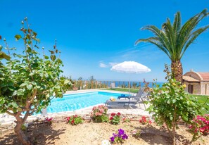 A swimming pool with lounge chairs and a palm tree, surrounded by colorful flowers and a clear blue sky. - Villa Lygaria (Malevizi)