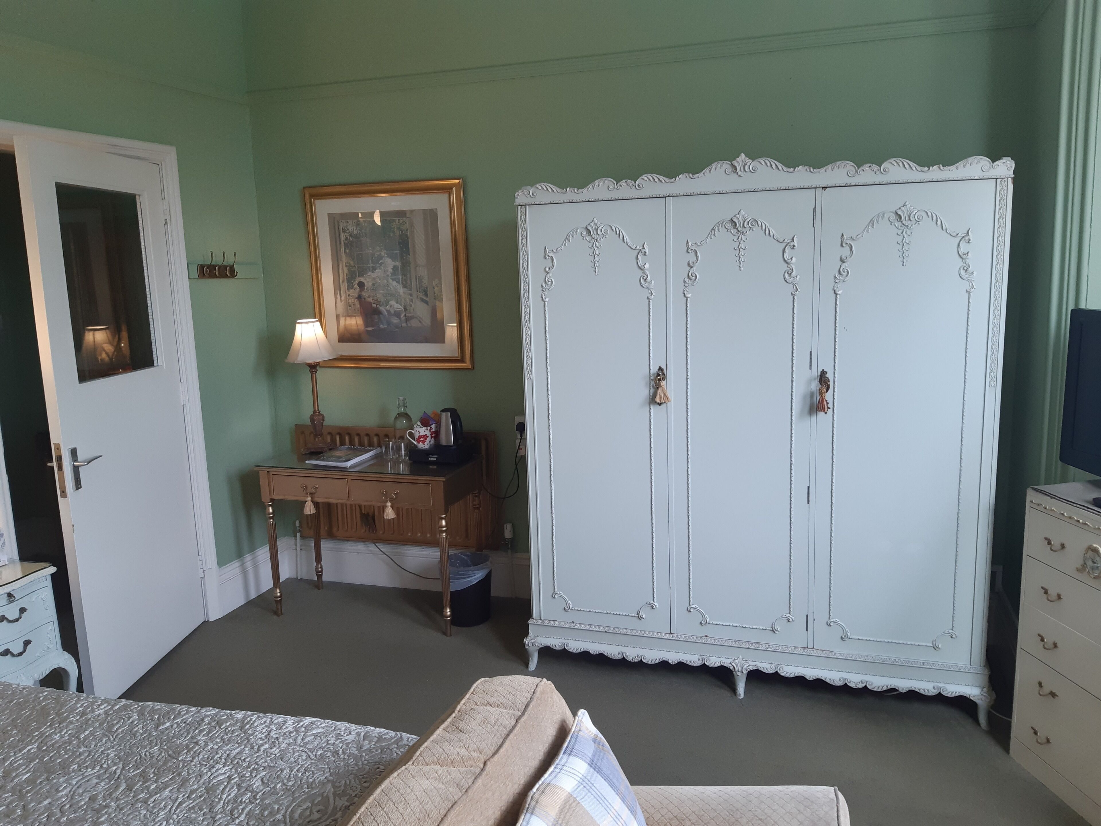 A room with a bed, a desk with a lamp, a wardrobe, and a painting on the wall.
