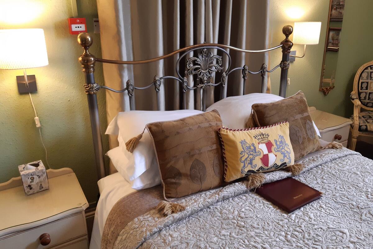 A neatly made bed with a patterned blanket and decorative pillows, a bedside table with a tissue box, and a wall-mounted lamp.