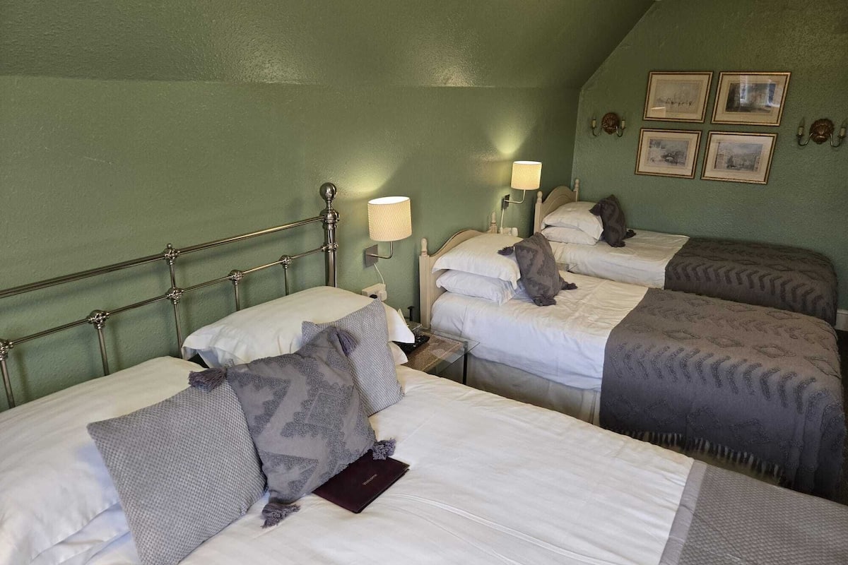 A room with three beds, each with a headboard and bedside lamps. There are framed pictures on the wall and a lamp on the nightstand.