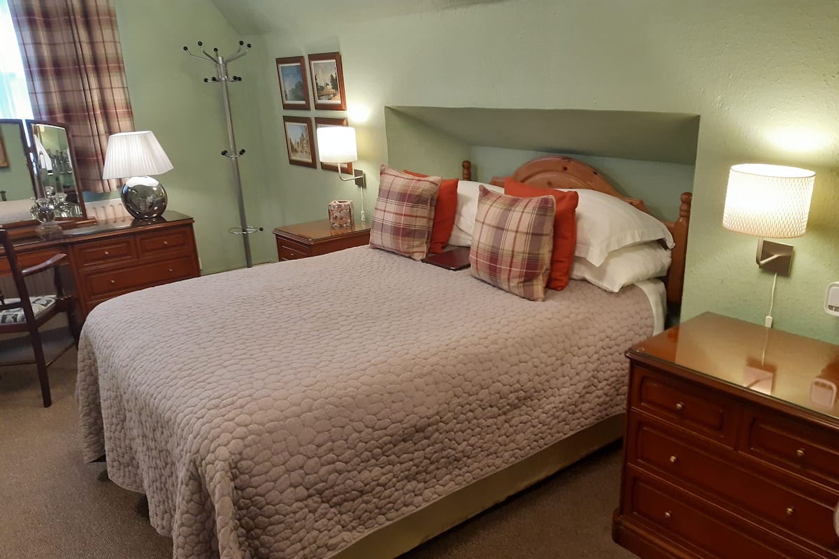 A bedroom with a large bed, wooden nightstands, a desk, and a chair. There are two wall-mounted lamps and a mirror.
