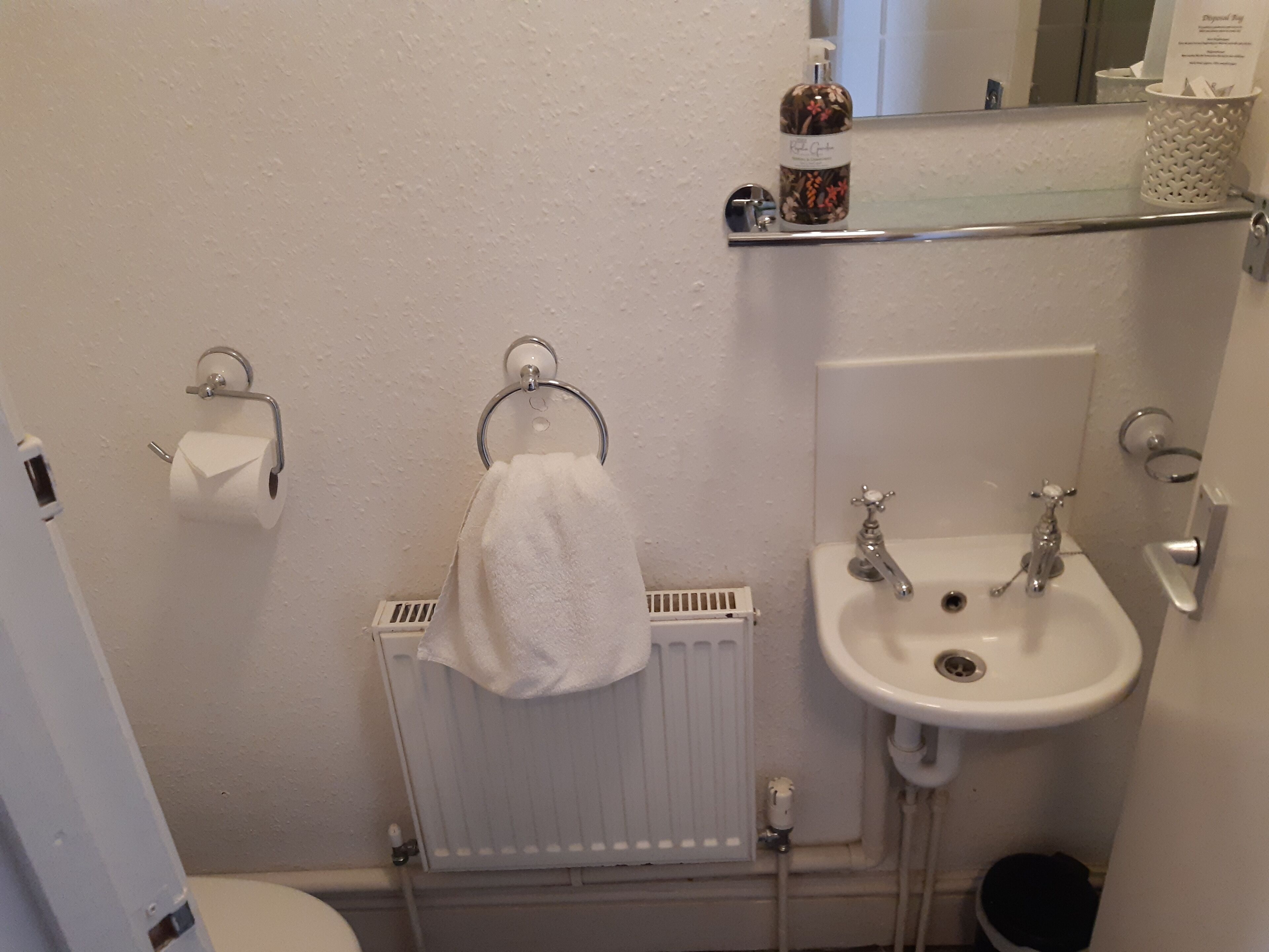Un baño con un lavabo blanco, un inodoro, un perchero para toallas, un dispensador de papel higiénico y un radiador.