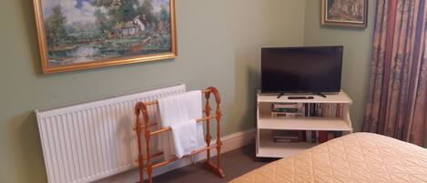 A bedroom with a bed, a radiator, a wooden towel rack, a television on a stand, and two framed pictures on the wall.