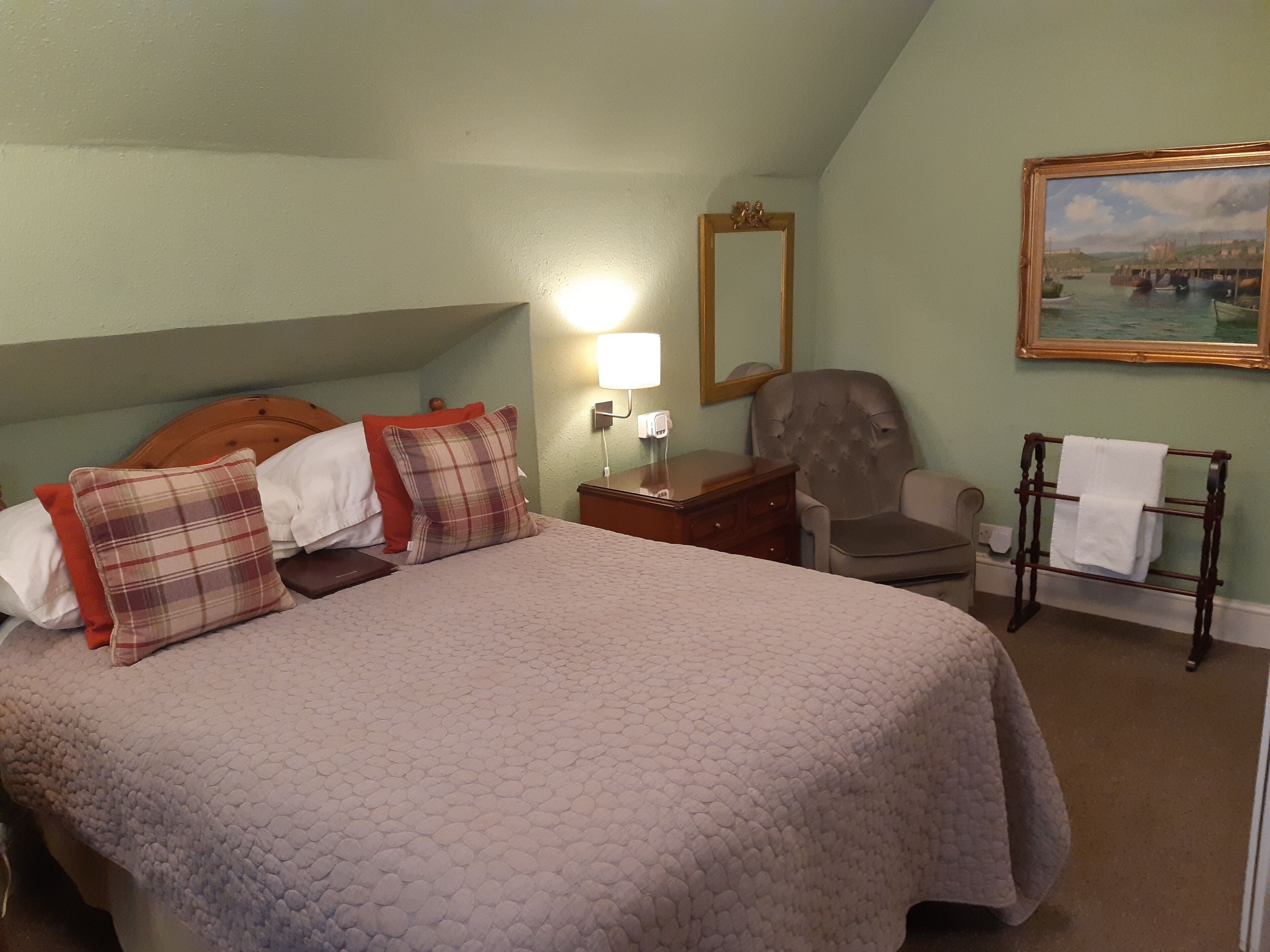 A neatly made bed with a quilted cover, a wooden headboard, and several pillows. There is a chair, a small table with a lamp, and a wall-mounted mirror. A painting of a harbor scene is visible on the wall.
