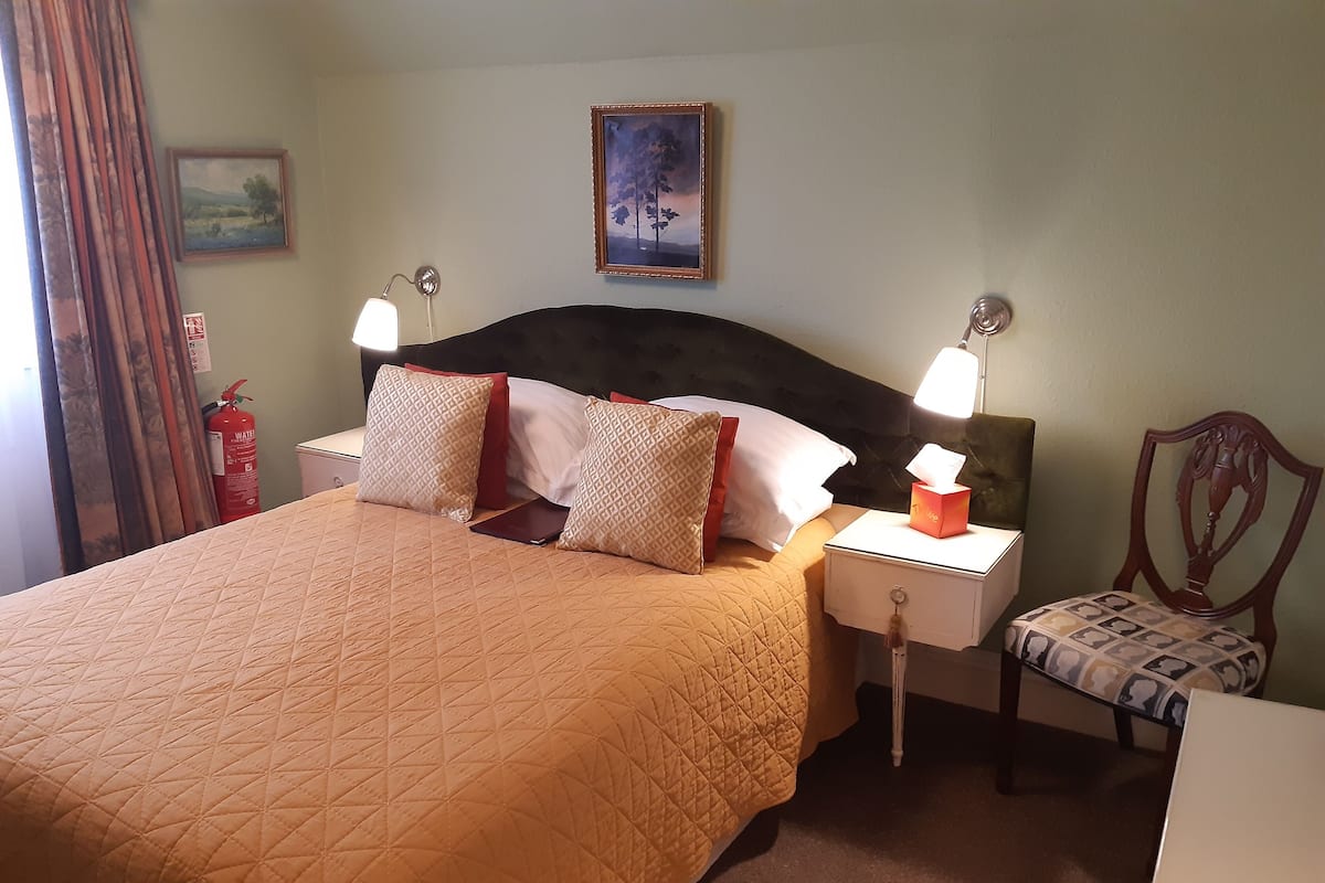 A hotel room with a bed, nightstands, a chair, a fire extinguisher, and a painting on the wall.