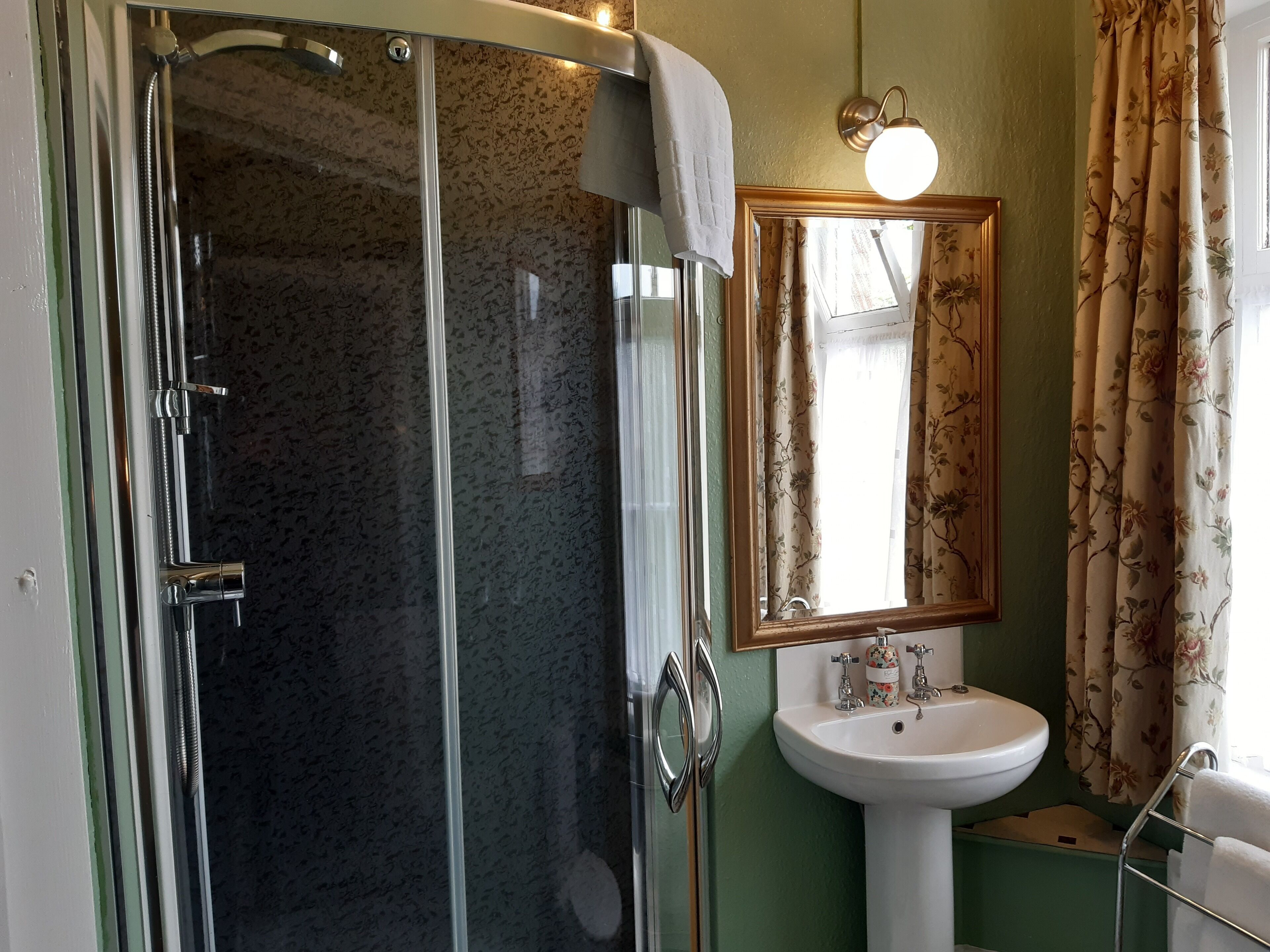 A bathroom with a glass-enclosed shower, a white sink, and a mirror with a floral curtain.