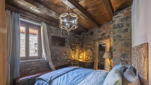Un dormitorio rústico con pared de piedra, techo de madera, cama con ropa de cama azul, escritorio con lámpara y televisor montado en la pared.