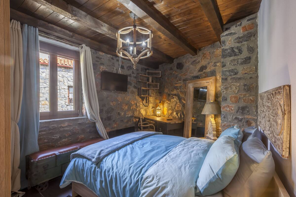 Un dormitorio rústico con pared de piedra, techo de madera, cama con ropa de cama azul, escritorio con lámpara y televisor montado en la pared.