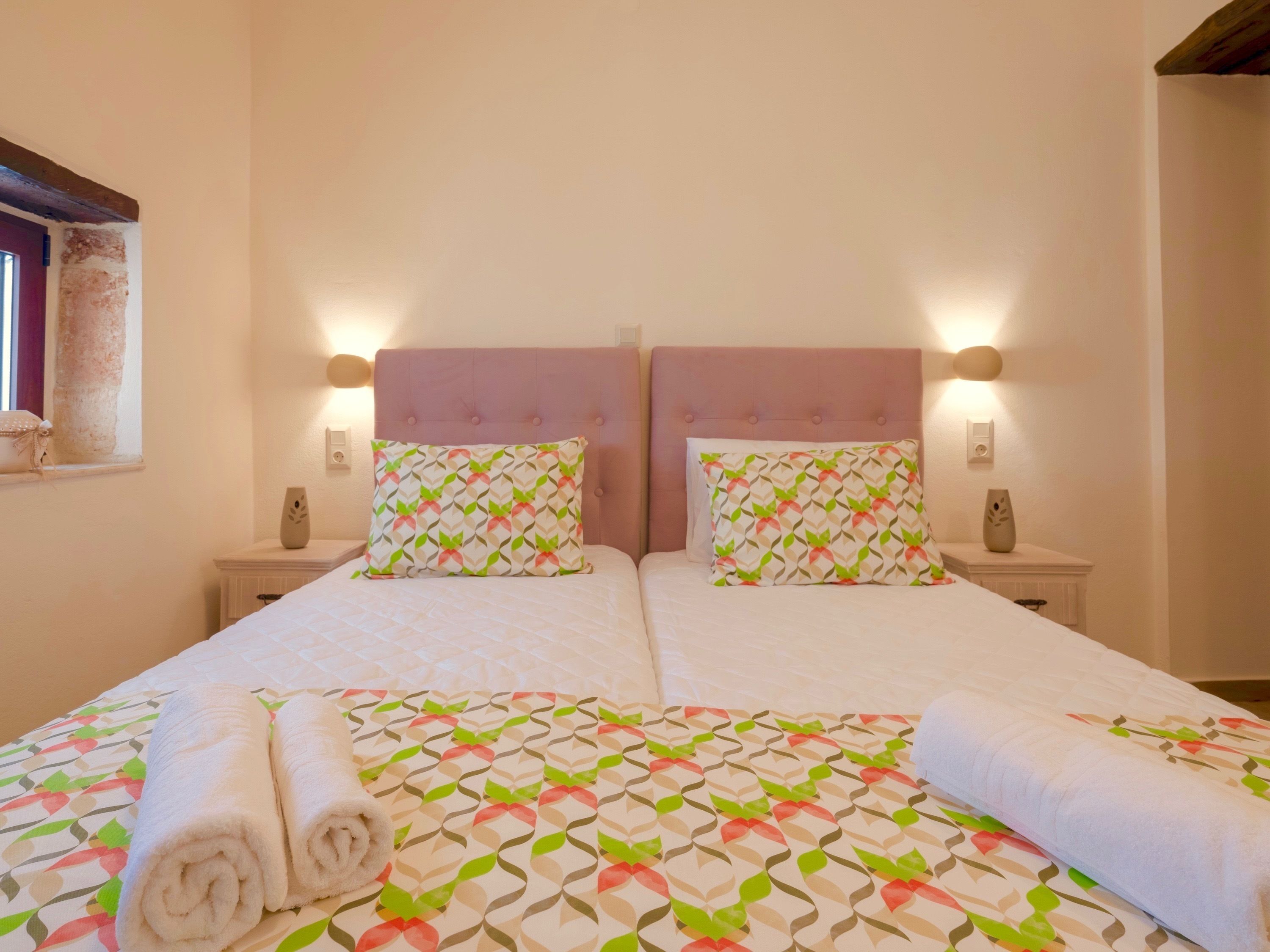 A double bed with a patterned bedspread and two matching pillows, a nightstand with a vase, and a window with a view of the outside.