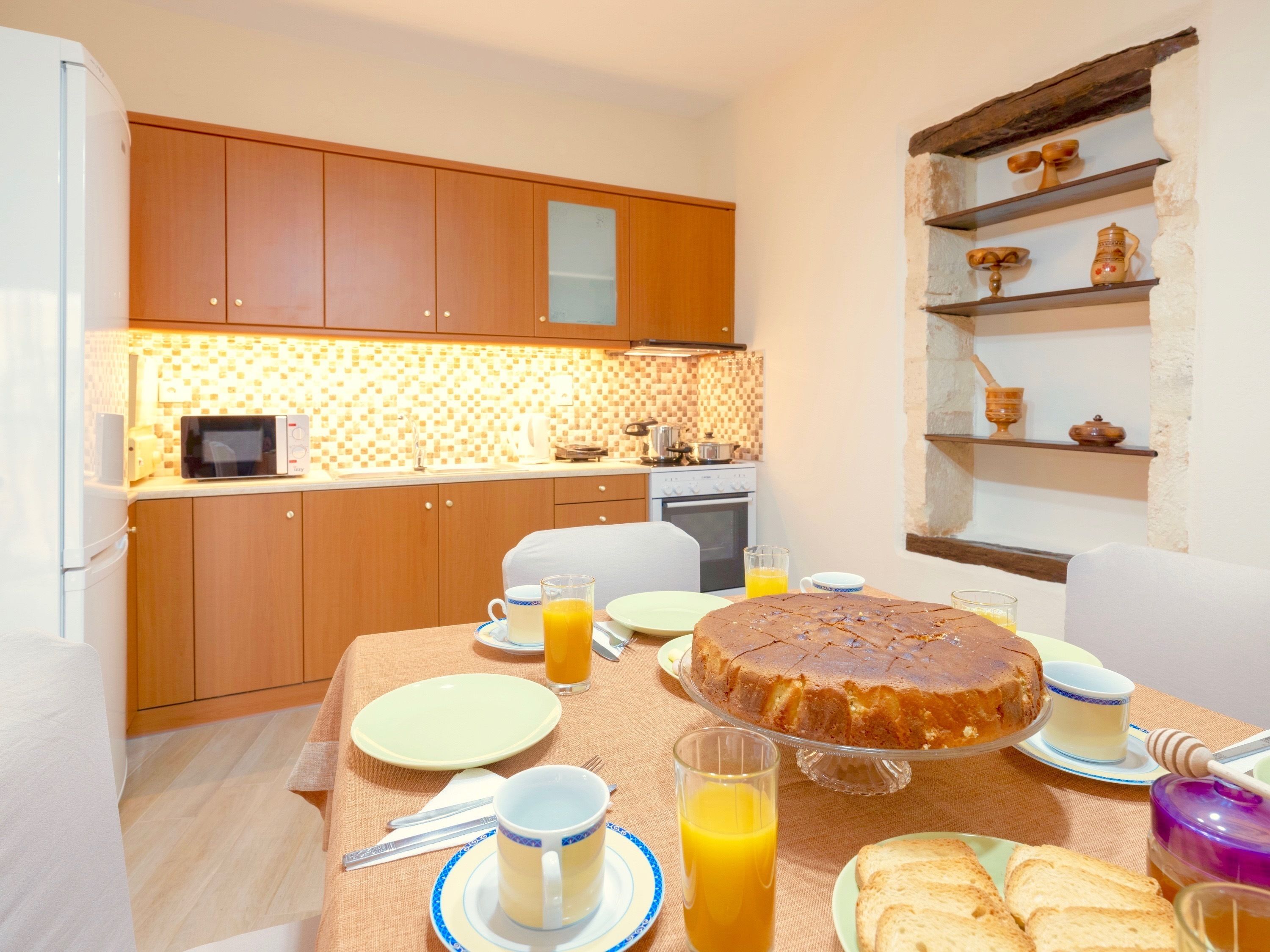 A breakfast setup with a cake, orange juice, and toast on a table in a kitchen with wooden cabinets and a tiled backsplash.