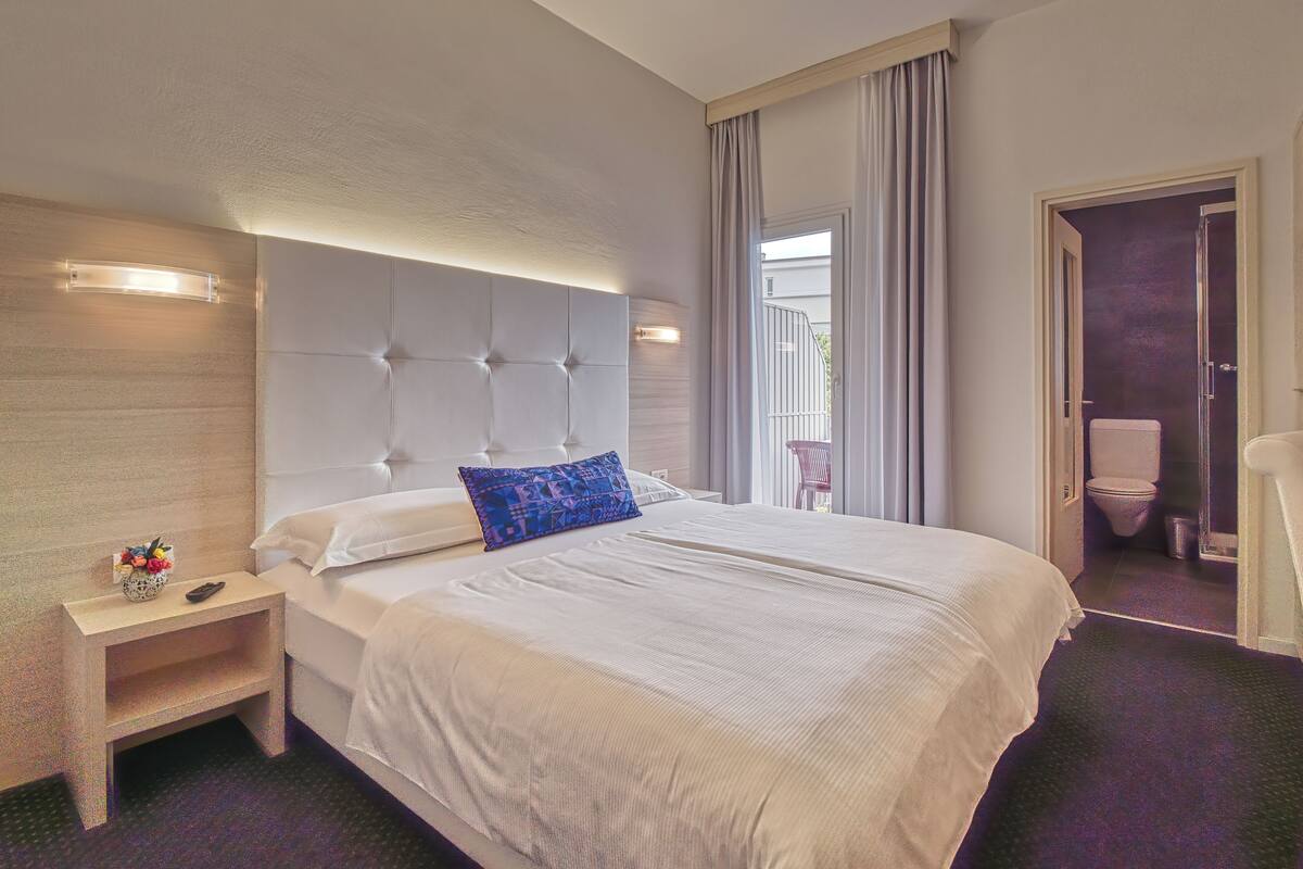 A hotel room with a large bed, a bedside table with a vase, and a view of the outside balcony.