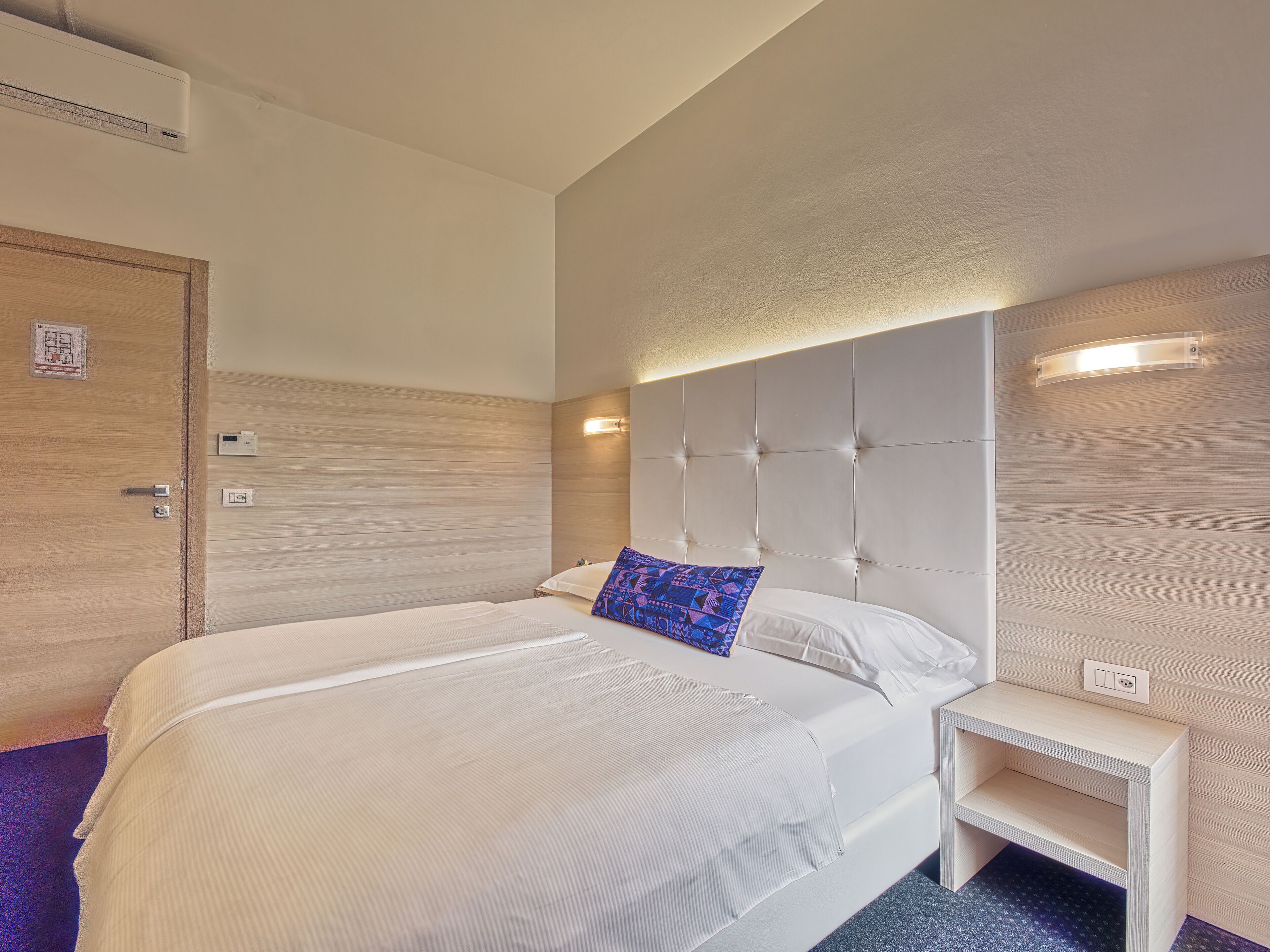 A hotel room with a large bed, a bedside table, a built-in headboard, and a wall-mounted light fixture.