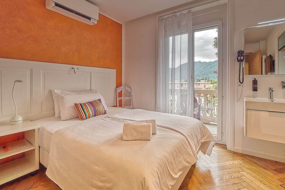 A hotel room with a bed, a bedside table, a white air conditioner, a bathroom with a sink and mirror, and a balcony with a view of mountains.