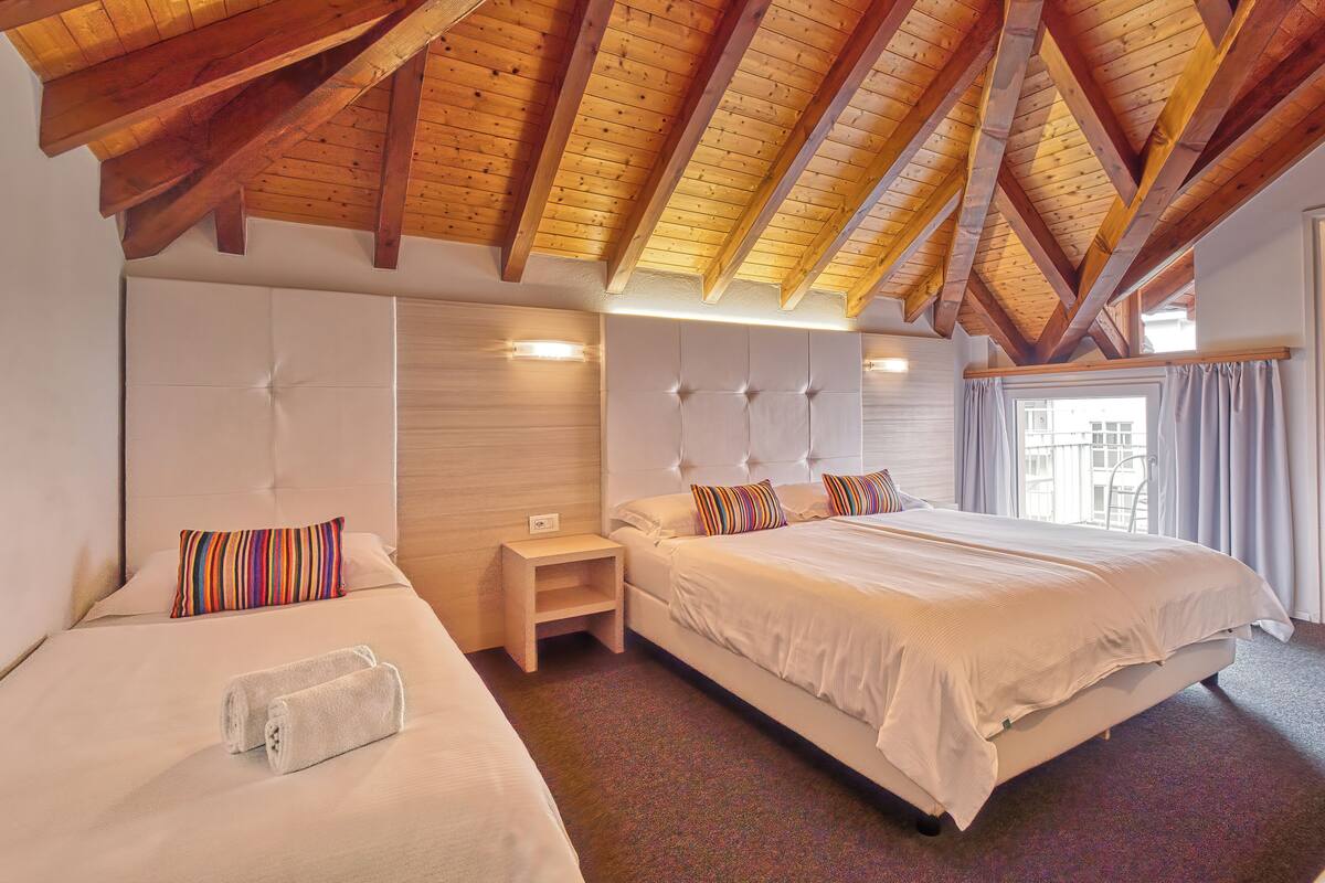 A hotel room with two beds, wooden beams on the ceiling, a large window with curtains, and a bedside table with a lamp.