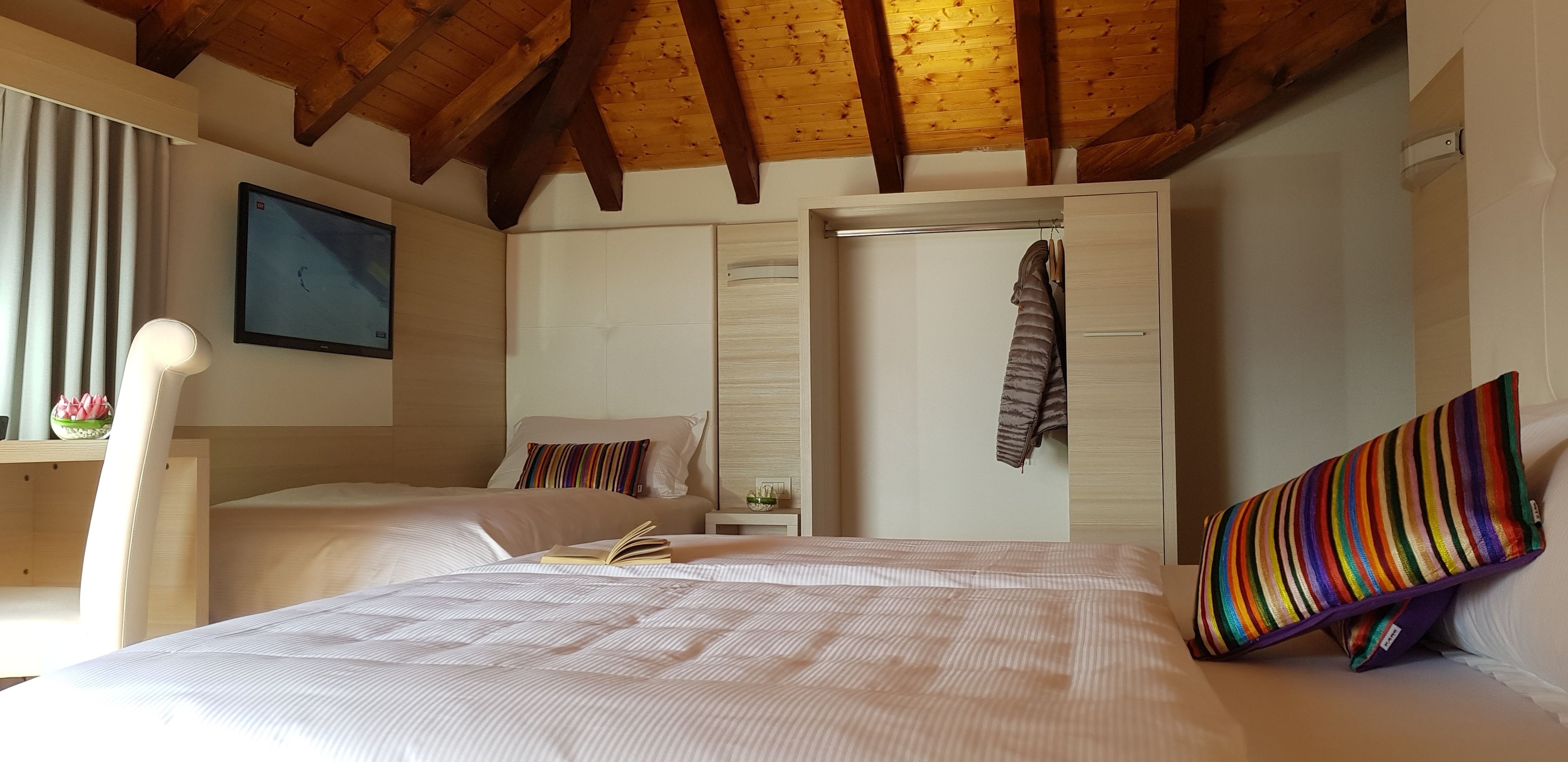 A bedroom with a wooden ceiling, a bed with a striped pillow, a bedside table with a lamp, and a TV on the wall.
