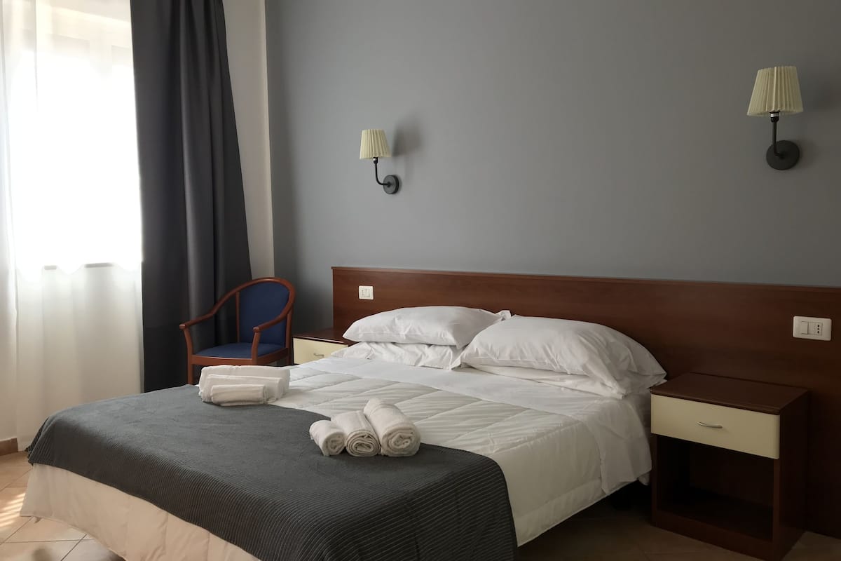 A neatly made bed with white linens, a dark grey blanket, and rolled towels on the foot of the bed. There is a wooden nightstand with a lamp, a chair, and a wall-mounted light fixture.