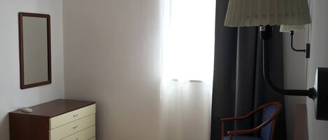 A hotel room with a bed, a dresser, a chair, a desk lamp, and an air conditioning unit.