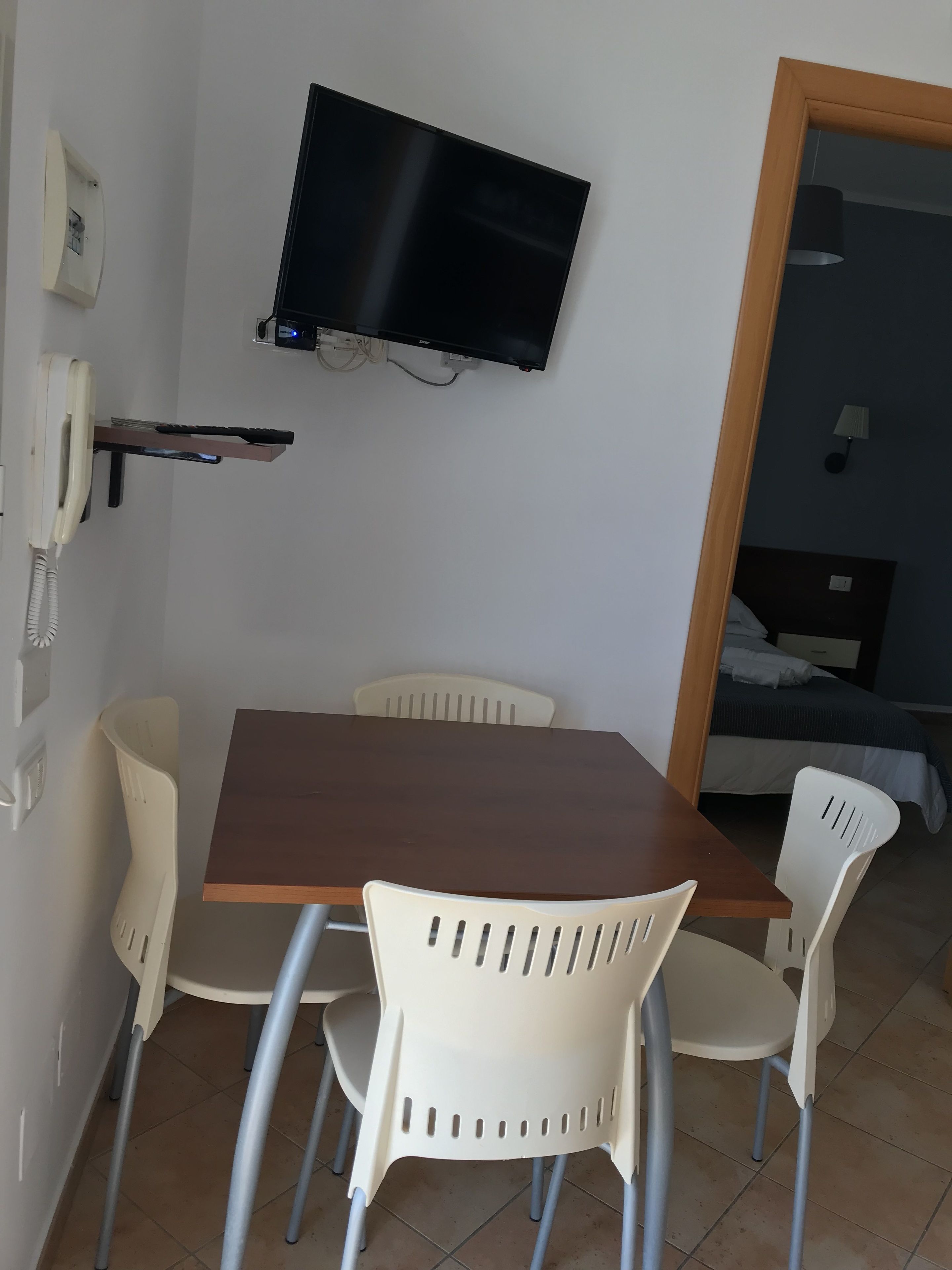 Une petite pièce avec une table en bois et quatre chaises en plastique blanc. Un téléviseur à écran plat est fixé au mur, au-dessus de la table.
