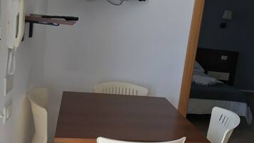 Une petite pièce avec une table en bois et quatre chaises en plastique blanc. Un téléviseur à écran plat est fixé au mur, au-dessus de la table.