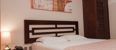 A hotel room with a wooden headboard, a bed with white linens, a patterned throw blanket, a nightstand with a lamp and telephone, and a wall-mounted abstract painting.