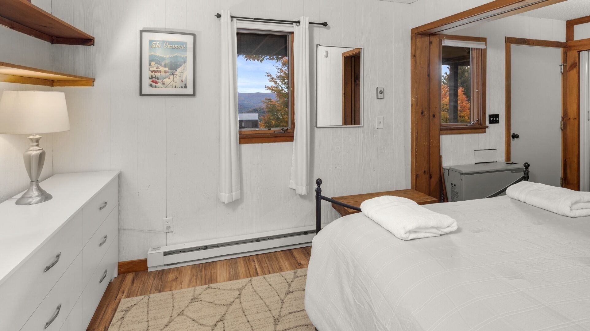 Un dormitorio con cama, mesita de noche con lámpara, calefactor, ventana con cortinas y un cuadro en la pared.