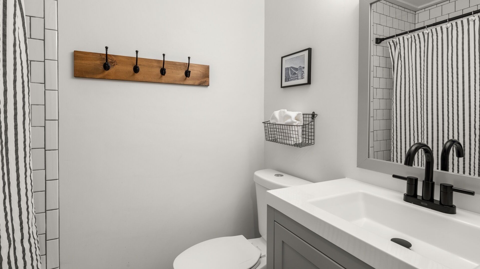 A bathroom with a white toilet, a white sink with a black faucet, a wooden wall-mounted rack with hooks, a framed picture, and a towel rack with a white towel.