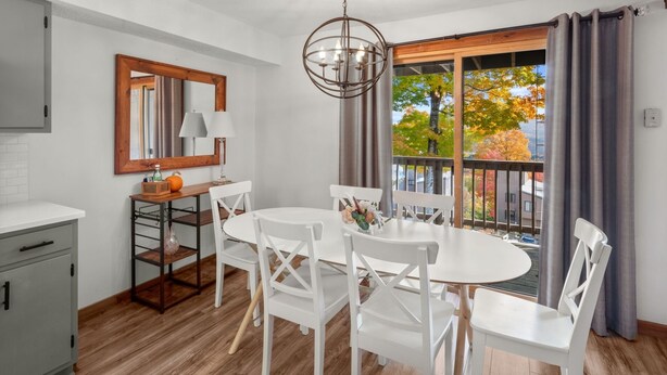 A dining area with a round table, white chairs, a chandelier, a wooden cabinet, and a window with curtains. - 57 North Lynx 3 Bedroom Condo by RedAwning (Warren)