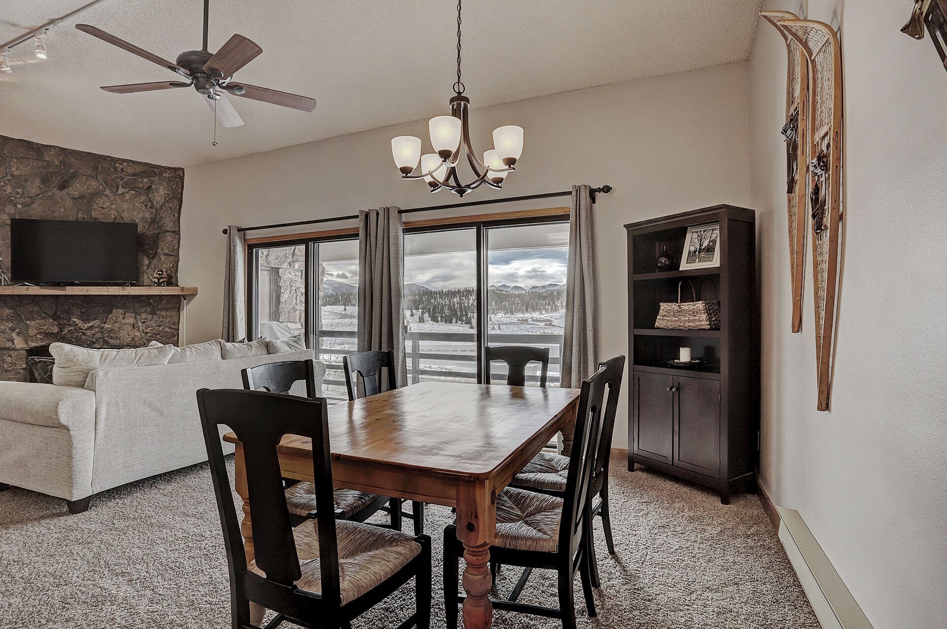 A dining area with a wooden table and chairs, a ceiling fan, a chandelier, and a view of a snowy landscape through large windows.