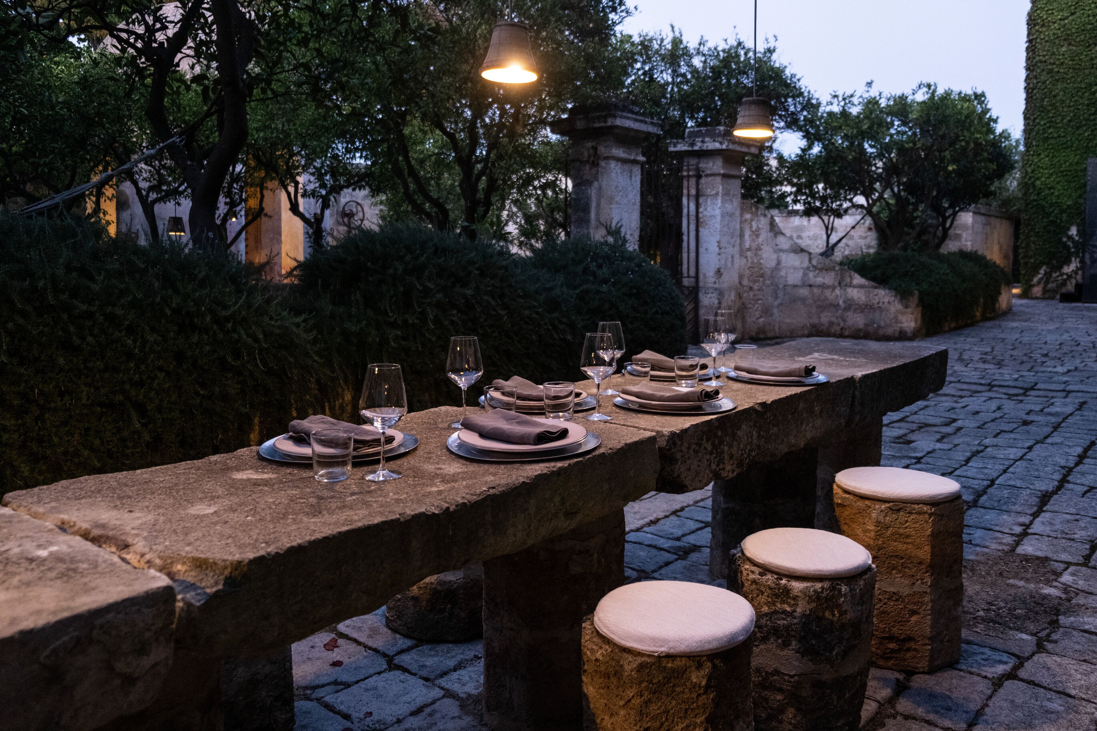 Ein Steintisch mit Tellern, Gläsern und Servietten, umgeben von Stehstühlen, in einer Gartenanlage mit Steinmauern und Grünpflanzen.