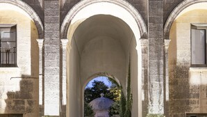 Un couloir bordé d’arches mène à une cour intérieure où se trouve une fontaine, et un bâtiment se dresse à l’arrière-plan.